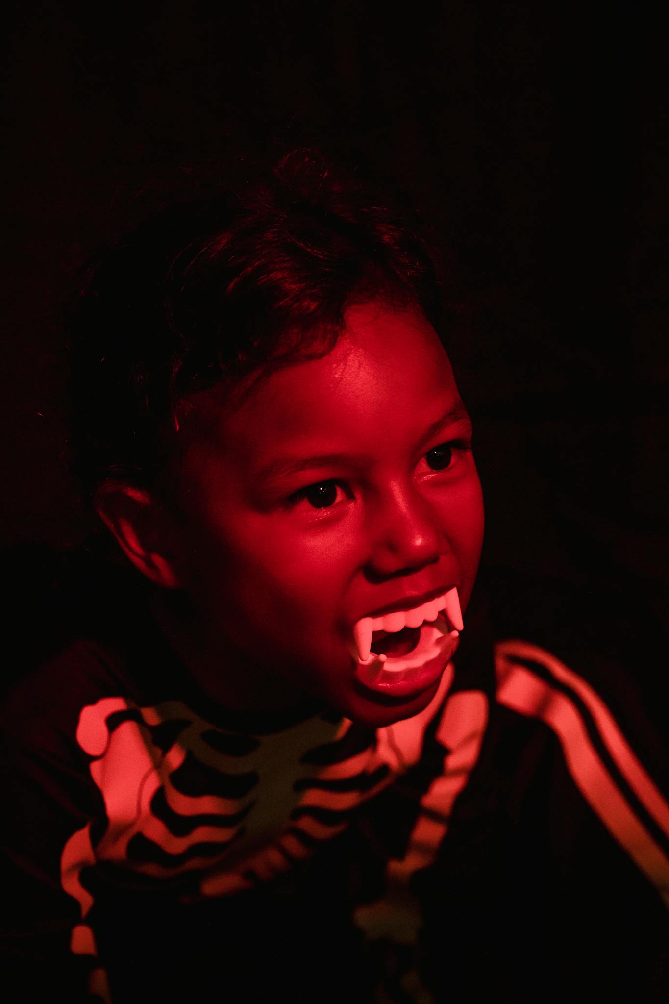 A child in a skeleton costume wearing vampire teeth against a dark background.